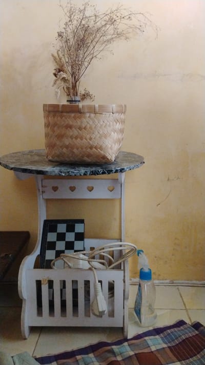 A wooden basket filled with dried plants sits on a round table with a marble-like surface. The table is supported by a white wooden stand with heart cutouts. Below the table, a portable chessboard is placed in a white wooden holder. A white electrical cord with a plug is draped over the holder. To the right of the holder, a clear plastic spray bottle with a blue cap stands upright. The background consists of a textured yellow wall and tiled flooring. A checkered fabric lies on the floor in the foreground. The scene is indoors, likely in a residential setting within Banjarsari, Indonesia.