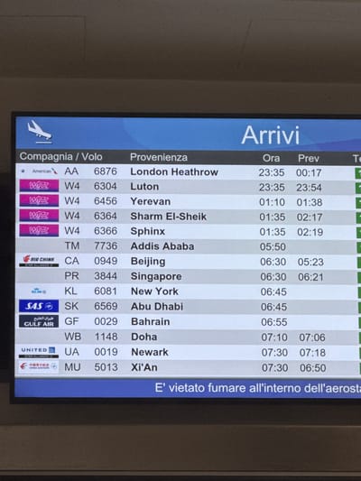 This is a photograph of an airport arrivals board displaying flight information. The board is primarily blue with white and grey text. The text is oriented vertically, suggesting the photo was taken with the camera rotated 90 degrees.
The board lists the airline ("Compagnia / Volo"), the origin of the flight ("Provenienza"), the scheduled arrival time ("Ora"), and the previous scheduled arrival time ("Prev"). There is also a column with what appears to be gate information ("Te"), indicated by numbers.
Several airlines are visible, including American Airlines, Wizz Air, Air China, KLM, SAS, Gulf Air, and United. The destinations listed are London Heathrow, Luton, Yerevan, Sharm El-Sheik, Sphinx, Addis Ababa, Beijing, Singapore, New York, Abu Dhabi, Bahrain, Doha, Newark, and Xi'An. The times range from late evening (23:35) to early morning (07:30).
A notable piece of text at the bottom right reads "E' vietato fumare all'interno dell'aerosta," which translates to "Smoking is prohibited inside the airport."
The scene appears to be inside an airport terminal. The lighting suggests it could be nighttime or early morning, given the flight times. There are no people clearly visible in the image, only the information displayed on the board. The overall impression is functional and informative, characteristic of an airport information display.