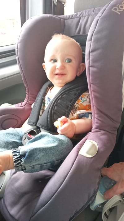 The image shows a baby in a car seat, likely in a vehicle. The baby, with blonde hair and blue eyes, is looking towards the right of the frame with a slightly surprised or amused expression. The baby is wearing a colorful patterned shirt and denim jeans. The car seat is a deep purple color and has black harness straps. The baby's left hand is holding a small, orange object, possibly a piece of food. A hand is visible on the right side of the frame, holding a blue and white baby bottle. The scene appears to be indoors, possibly during the daytime, with light coming from the left, suggesting a window. There is no visible text in the image that provides context. The location is Altdorf bei Nürnberg, Germany.
