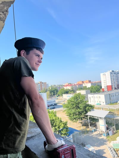 A young man wearing a dark, brimmed cap and an olive green t-shirt stands on a concrete structure, looking out over a cityscape under a clear blue sky. His left arm is resting on the concrete ledge, and he is holding a red power tool in his right hand, which is covered by a white glove. The background shows a mix of residential and industrial buildings, with trees and parked cars visible. The image appears to be taken from a high vantage point, possibly a balcony or rooftop, suggesting an urban or semi-urban setting. The specific location is identified as Аснежыцкі сельскі Савет, Belarus.