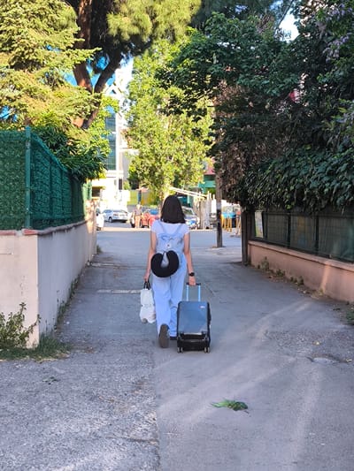 A young woman walks away from the camera down a narrow street, pulling a black wheeled suitcase behind her. She is wearing a light-colored jumpsuit and a light blue backpack. A black U-shaped travel pillow is attached to the backpack's straps. She is also carrying a white shopping bag in her left hand. The street is paved with asphalt and lined with a low stone wall and a green fence on the left, and a pinkish wall with a wire fence on the right. Lush green trees and foliage overhang the street from both sides, casting dappled shadows.
In the background, several cars are parked along the street, including a white sedan and a red sedan. There are also buildings visible, some of which appear to be residential or commercial. The scene suggests an urban or semi-urban environment. The lighting indicates it is daytime, likely late afternoon due to the warm light and long shadows. The presence of the travel pillow and suitcase suggests the woman might be traveling, perhaps to or from a train station, bus stop, or airport, or simply moving locations.
A pair of green parrots are visible on the asphalt in the foreground, near the right edge of the frame. They appear to be feeding or resting. There is some visible text on the white shopping bag, but it is not legible. The overall mood is calm and ordinary, capturing a moment of everyday movement.