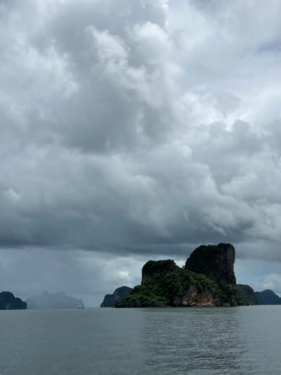 The image shows a body of water with several rocky islands covered in vegetation. The sky is overcast with dramatic, grey clouds. In the foreground, the water is calm with gentle ripples. The main subject is a large, imposing island in the middle ground, characterized by a sheer rock face and a lush green canopy of trees. To the left and right, smaller islands are visible in the distance, some appearing hazy. A small boat is present in the far distance between two islands. The scene evokes a sense of natural beauty and perhaps a hint of foreboding due to the stormy sky. This landscape is characteristic of the limestone karsts found in Krabi province, Thailand, which includes the region of Krasom.