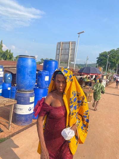 This image depicts a bright outdoor street scene in Amaseri, Nigeria, under a clear blue sky with scattered white clouds. The foreground is occupied by a woman walking leftward, her right profile visible. She wears a dark red, off-shoulder sequined dress and a vibrant yellow headwrap patterned with blue and black geometric shapes. Her face shows a neutral expression, with eyes cast downwards. Her visible right arm appears slender and wrinkled, and she holds a small, crumpled white cloth object in her left hand, partially obscured by her headwrap.
To the woman's left, multiple blue plastic industrial barrels are stacked on an unpaved, light brown earth surface. One barrel in the lower-left foreground displays a white label with "RAJ PETRO" text and a logo. A small wooden table adjacent to the barrels holds a scattering of nuts, consistent with peanuts. The woman and the running child are on a reddish-brown paved road that extends into the background.
In the midground, a child wearing a green patterned tunic and flip-flops runs rightward into the background. Behind the child, the scene transitions into a bustling street market, characterized by several stalls featuring dark blue umbrellas providing shade. Various goods are visible on tables at these stalls, with other individuals discernible among them.
The background includes urban infrastructure such as a large, metal-paneled sign structure and multiple tall streetlights, one of which is a solar-powered unit. Lush green trees line the horizon, beneath which distant hills are visible. The scene is illuminated by bright natural light, casting distinct shadows.