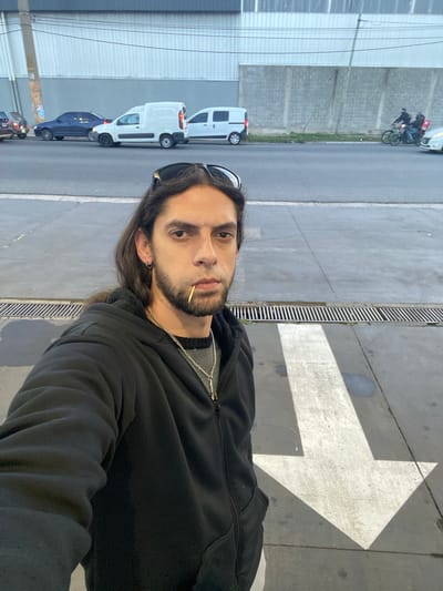 A medium-shot photograph of a man with long dark hair, wearing sunglasses and a black hooded jacket with a silver chain around his neck. He has a toothpick in his mouth. The photograph is taken on a street with parked cars and a motorcycle in the background. A large arrow is painted on the sidewalk. The location is in Quilmes, Argentina.