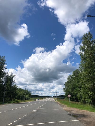 A wide, paved road stretches into the distance, curving gently upwards. The road is lined on both sides by lush green trees and foliage. The sky above is a vibrant blue, dotted with dramatic, fluffy white and grey clouds. A few streetlights are visible along the roadside. In the distance, a dark-colored car is seen driving on the road. A circular traffic sign with a red border and a white background, featuring two red cars inside, is positioned on the right side of the road. The sign appears to indicate a prohibition for certain types of vehicles. The overall scene depicts a scenic view of a road in a natural setting, under a dynamic sky. The location is Vysotnik, Russia.