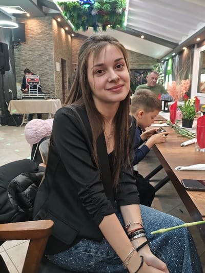 The image depicts the interior of a dining or event establishment in Opaka, Bulgaria. A young adult female is seated in the foreground, slightly off-center, facing the camera with a subtle smile. She has long, straight brown hair with bangs, wears a black blazer over a dark top, and blue jeans with small white speckles. A silver-colored necklace with a pendant and multiple bracelets (black, silver-toned, red string) are visible. A black puffer jacket rests on the chair to her left.

To her right, a young male child is seated at a dark wooden table, looking down at a small dark electronic device. Further back and slightly right, an older adult male in a green long-sleeved shirt is seated at a table. The wooden table in the foreground is set with red napkins, glasses, and small decorative plants; a dark-cased smartphone lies on its surface.

In the left background, a person operates a DJ setup consisting of turntables and a mixer on a draped table, accompanied by a large black speaker on a stand. The walls are textured with gray stone-like tiles. The ceiling is white with dark beams, recessed lighting, and suspended decorative greenery with some pink elements. The floor is light-toned tiled, reflecting some light.
