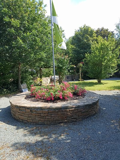 A circular, stone-lined flower bed filled with pink roses is the focal point of this outdoor scene. A flagpole with a green and white flag stands behind the flower bed. The background features lush green trees and a grassy area. A stone monument with an inscription is visible to the left of the flower bed. The ground surrounding the flower bed is covered in small grey gravel. The location is an outdoor public space, possibly a park or memorial area in Dungloe, Ireland.