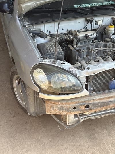 The image shows the front section of a damaged silver car with its hood open, revealing the engine compartment. The car is parked on a dusty, unpaved surface, likely a roadside or a yard. The front bumper is heavily rusted and detached, exposing the car's internal components, including the radiator. The left headlight is intact but the surrounding body panel appears to be slightly misaligned. The tire on the left front is visible and appears to be dirty. The overall condition of the car suggests it has been in an accident or is undergoing repairs. The environment appears to be an outdoor setting in a rural or semi-urban area. There are no people or animals visible in the image. The license plate, partially visible at the top of the windshield, is from Kyrgyzstan.