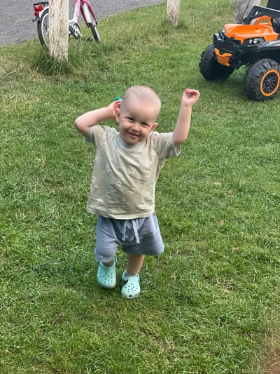 A young child, appearing to be a toddler, stands on a grassy area in Beltershain, Germany. The child is wearing a light-colored t-shirt with a dragonfly pattern and light blue shorts. They have on light blue Croc-style shoes. The child has their right arm raised and their head tilted back slightly, with a wide smile on their face. In the background, to the left, a pink bicycle is leaning against a white post. To the right, a bright orange and black ride-on toy vehicle is visible. The ground is covered in green grass, and in the distance, there are hints of trees and other vegetation.