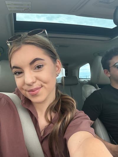 A medium shot captures two individuals inside a vehicle, with a woman in the foreground and a man partially visible in the background. The woman, positioned on the left, is looking towards the camera with a slight smile. She has light brown hair pulled back, wears a brown collared shirt, and a beige seatbelt is across her chest. Sunglasses are perched on her head. To her right, the man is seated, facing forward, wearing a black t-shirt and sunglasses. The interior of the car is visible, including the sunroof and grey upholstery. The overall scene suggests an outdoor setting, possibly during daytime. The exact location of Aldomirovtsi, Bulgaria, cannot be determined from the visual information alone.