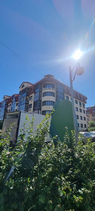 The image captures a bright, sunny day in Romashkovo, Russia, with the sun prominently featured in the upper left quadrant, casting a lens flare across the scene. The primary subject is a modern, multi-story building with a distinctive facade featuring large windows and some brickwork. In front of the building, a lush green bush occupies the right side of the frame, its branches reaching upwards and partially obscuring the building.

To the left of the building, a street lamp with an unusual, spherical light fixture stands tall. The sky is a clear, vibrant blue. The building itself appears to be residential, with visible air conditioning units on several floors, suggesting a warm climate or season. The architecture is contemporary, with a mix of glass, lighter-colored brick, and darker rooflines. A portion of another building, with a similar architectural style, is visible in the background.

The overall impression is one of an urban or suburban residential area on a pleasant day. The sunlight suggests it is daytime, likely late morning or early afternoon given the intensity of the sun and the clear sky, indicating fair weather. No people are visible in the image, and there is no visible text. The scene conveys a sense of calm and ordinariness, typical of a residential neighborhood.