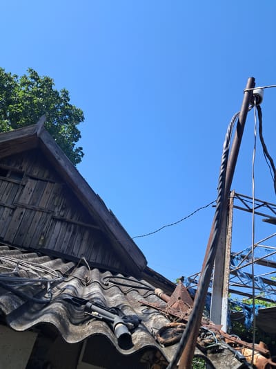 This media file captures an upward-angle view of an outdoor scene under a clear blue sky. The foreground is dominated by a rustic wooden structure, possibly part of a building or shed, with weathered planks and some exposed framing. On top of this structure, a section of corrugated roofing is visible, along with various electrical wires and a rusty metal pole extending upwards. A tangled mass of wires and metal supports suggests an abandoned or disused piece of infrastructure, possibly related to power lines or an antenna. In the background, a lush green tree with dense foliage is partially visible, contrasting with the decaying man-made elements.

The time of day appears to be daytime, indicated by the bright blue sky and the presence of sunlight casting shadows. The weather seems fair and clear. The scene evokes a sense of disrepair and abandonment, with the rusted metal and weathered wood hinting at neglect. The setting could be a rural or semi-rural area, given the natural elements like the tree and the rustic construction. There are no people visible, and the focus is on the objects and the environment. No text is discernible within the image. The overall impression is one of decay juxtaposed with the enduring presence of nature.