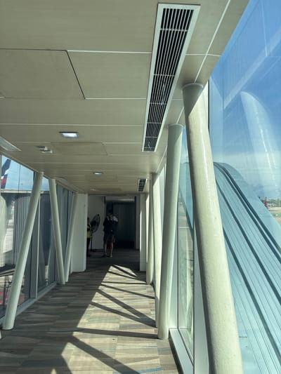 This image depicts the interior of a jet bridge or airport walkway at Phuket International Airport (HKT) in Mai Khao, Thailand. The perspective is from within the walkway, looking towards its far end.
The ceiling consists of light-colored rectangular panels, some showing minor discoloration or misalignment. Integrated into the ceiling are several rectangular recessed light fixtures and long, gridded rectangular air conditioning vents.
The floor is carpeted with a patterned design composed of horizontal stripes in varying shades of grey, blue, and beige. Direct sunlight streams through the windows on the right, casting prominent, elongated diagonal shadows across the floor.
Along the right side of the walkway, a series of thick, cylindrical support columns, painted a light, textured off-white color, are angled inward from the base to the ceiling. These columns stand between a full-height glass window wall that runs the length of the walkway.
Through the window, an external airport environment is visible. A large, curved architectural structure with a ribbed or corrugated metallic surface, possibly a roof or part of another terminal building, occupies a significant portion of the outdoor view. Beyond this structure, a blue sky with some cloud cover is apparent. Further in the distance, parts of a red and white aircraft, along with a light pole, are discernible on the tarmac.
Towards the far end of the walkway, two individuals are present. One individual, closer to the center, is partially obscured but appears to be wearing a high-visibility vest and is near a black portable fan positioned on the floor. The second individual, further down the walkway, is seen from behind, wearing a dark hooded top, shorts, and a backpack, and appears to be standing or walking away from the viewer. The far end of the walkway leads to an interior area with a solid wall.