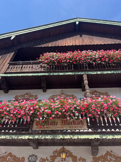 The image displays the upper facade of Gasthütte Marbach, located at Marbach 39, Fischbachau, Germany, under a clear blue sky. The building exhibits traditional Bavarian architectural features, including dark brown wooden siding on the upper story and white stucco on the lower visible sections. Two wooden balconies, each with an intricately carved balustrade, span the facade. Both balconies are extensively adorned with dense arrangements of flowering plants, primarily pink and red geraniums, which overflow from flower boxes. A rectangular wooden sign is mounted on the white stucco wall behind the lower balcony's flowers. The sign reads "Gasthütte Marbach" in a Gothic script, with smaller text below it stating "FREMDENZIMMER," "ANTIK STUBE," and "EIGENE METZGEREI." The white stucco wall sections feature ornate, gold-toned painted scrollwork and floral designs. A circular plaque bearing the number "39" is visible on the lower left portion of the stucco wall, alongside a single hanging lantern-style light fixture. No individuals, actions, or interactions are present in the scene.
