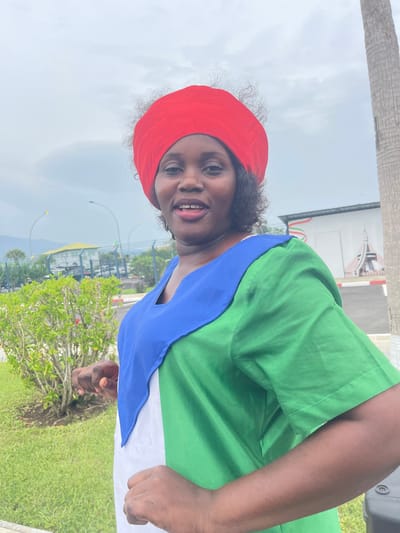 An outdoor portrait shot in Malabo, Equatorial Guinea, features a smiling woman. The image is oriented sideways, but if rotated correctly, the woman is positioned on the right side of the frame, looking towards the viewer with a broad smile, showing her teeth. She wears a bright red headscarf that covers her hair, and a green garment. A striking blue piece of fabric is draped over her chest and shoulders, with a section of white fabric visible beneath it. Her dark, curly hair is visible around the edges of the red head covering.

The background presents a blend of natural greenery and industrial elements under an overcast sky. Lush green grass and bushes with some reddish blooms are visible in the foreground and midground. To the woman's left, a segment of a palm tree trunk is present. In the upper right background, sections of industrial structures or buildings, possibly containing pipes and railings, can be discerned, suggesting an industrial or facility area. The sky is a uniform light gray, indicating a cloudy or overcast daytime. The scene conveys a sense of cheerfulness from the subject amidst a practical, outdoor setting. There is no visible text in the image.