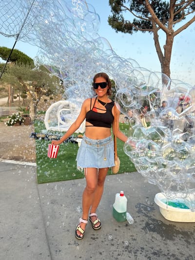 This is a full-shot, vertically oriented image of a woman creating a large cluster of bubbles outdoors. She is wearing a black crop top, a denim skirt, and sandals. Her body is angled towards the right of the frame, and she is looking at the camera with a smile. She is holding a wand with a large bubble net, which is producing the voluminous collection of bubbles that dominate the left side of the image. In her right hand, she holds a small, red-and-white striped container, possibly for popcorn or snacks.
The setting appears to be a sunny day with a clear blue sky. There is a large tree with branches extending from the top left of the frame. In the background, there is a patch of artificial turf and a white structure, possibly a tub or fountain, containing green water. A large bottle of green liquid, likely bubble solution, lies on the concrete ground to the right, with a small bottle on top.
The image conveys a sense of fun and playfulness, likely at a fair, festival, or outdoor event. The abundance of bubbles and the woman's cheerful expression contribute to this atmosphere. The lighting suggests it is daytime, possibly late afternoon, given the warm tones. The location cues of greenery and outdoor structures, combined with the context of Sant Vicenç De Montalt, Spain, suggest a pleasant, possibly Mediterranean environment. There is no visible text in the image.