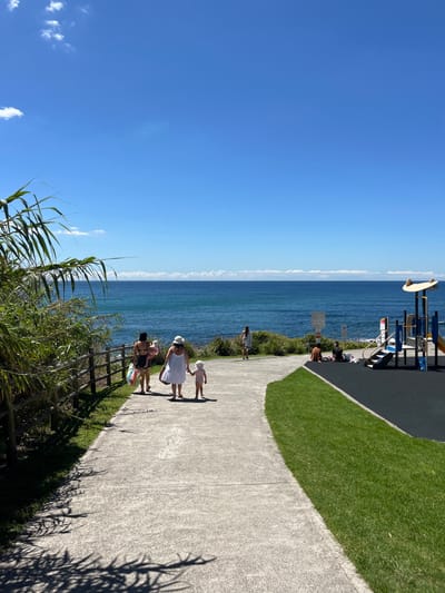 A coastal scene shows a paved pathway alongside the ocean. Several people walk along the path, including a woman in a white dress and hat holding a child's hand. Lush green grass and vegetation line the pathway. In the background is the deep blue ocean meeting a clear blue sky. A playground structure is visible in the upper portion of the image. A wooden fence and tall reeds are in the foreground.