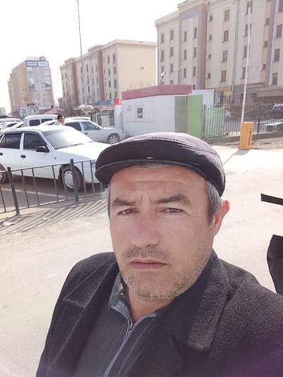 A middle-aged man with graying dark hair and stubble, wearing a dark flat cap, a dark overcoat, and a dark collared shirt, is captured in a selfie, looking directly at the camera with a neutral expression. The scene is an urban street environment in Urgench, Uzbekistan. Behind the man, a black metal barrier separates the foreground from a paved street. A queue of vehicles, including a prominent white Daewoo Nexia sedan and a white van, occupies the street, indicating parked or slow-moving traffic. In the midground, a white modular building with a red roofline, likely a kiosk, stands adjacent to a smaller green structure. The background features multiple multi-story residential or commercial buildings with light-colored facades, numerous windows, and decorative blue vertical accents. A tall light pole extends vertically on the left, and a yellow parking barrier arm is visible on the right. A large billboard or building-mounted sign displaying "EMS" is discernible on a distant building. The ground is paved, and the sky is bright and clear, consistent with daytime.