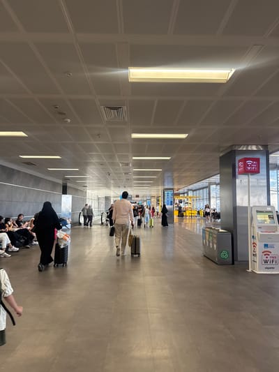 This is a vertical, eye-level, wide-angle shot of a modern, well-lit airport terminal in Pendik, Turkey. The ceiling is a grid of white panels with recessed rectangular lights running along the left side of the frame. The floor is a smooth, neutral-toned material.
The scene depicts a busy waiting area where travelers are gathered, some seated on the floor and others standing with their luggage. Several individuals are seen from behind, suggesting they are either waiting for a flight, a departure gate, or have just arrived. A group of people are seated on the floor on the right side of the frame, looking towards the left. Further down the terminal, people are standing in a line, presumably for check-in or security.
Notable objects include numerous suitcases and bags, some of which are black rolling suitcases, while others are duffel bags and shopping bags. Towards the upper part of the image, there are kiosks or information stands. One prominently displays "FREE WIFI" in red and white lettering, with accompanying logos. Another kiosk shows a screen with what appears to be flight information or a map. A gray recycling bin with green recycling symbols is also visible.
The lighting suggests it is daytime, with the artificial lights contributing to the bright and clean atmosphere of the terminal. There are no visible indications of weather conditions outside. The overall mood appears to be one of transit and anticipation, typical of an airport environment. The presence of "ISTANBUL" text on one of the signs provides a specific location cue.