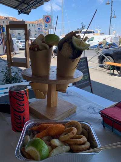 This image captures a close-up view of a food stall, likely in Porto Santo Stefano, Italy. The main subjects are two paper cones filled with fried seafood, each topped with a wedge of lime and a skewer. One cone is slightly obscured by the other, but both appear to contain a variety of fried items. To the right, a foil container holds more fried food, including onion rings and what look like shrimp. A can of Coca-Cola Zero Sugar is positioned in the foreground, its red and black branding clearly visible.
The setting appears to be outdoors, with a bright, sunny day indicated by the strong shadows and clear blue sky. In the background, a harbor with boats and buildings can be seen, along with a street scene featuring a vehicle and a parked scooter. A sign with Italian text, likely advertising the food, is visible behind the cones. The overall impression is of a casual, bustling atmosphere, perhaps a tourist area or a local market. There are no people prominently featured in the image, focusing the attention on the food and the setting. The time of day appears to be daytime, possibly late morning or afternoon, given the angle of the sun.