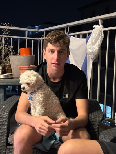 A man sits in a woven chair, holding a small, white, curly-haired dog. The man wears a black shirt and has short, curly brown hair. Behind him hangs a flag with blue stripes on a white background. A balcony railing is visible with plants below. Nighttime or a dark interior is suggested by the background darkness. Part of another person's leg is visible to the right.