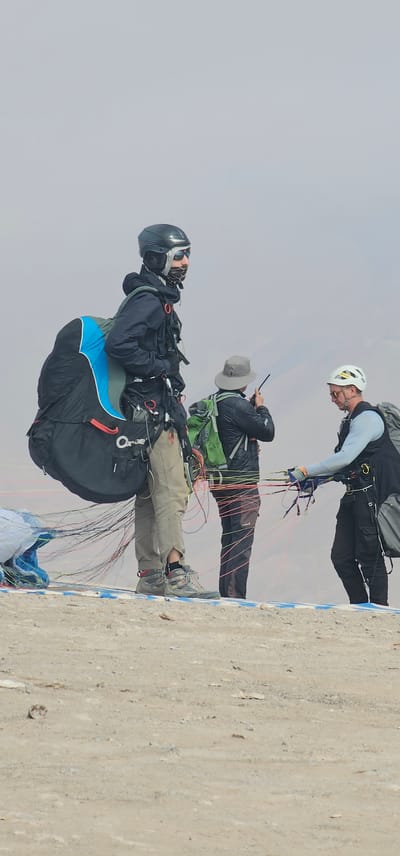 Three individuals are seen preparing for a paragliding session atop a concrete structure. The location is Alto Hospicio, Chile, given the arid, elevated landscape visible in the background. The leftmost person, wearing a dark helmet and goggles, is secured in a paragliding harness. The individual in the middle wears a hat and a dark jacket, along with a green backpack, and appears to be operating a device (likely a radio or smartphone). The person on the right, donning a white helmet, is checking lines connected to a paragliding wing. The concrete structure has a blue design on its edge. The environment is overcast with a grey sky.