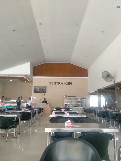 A brightly lit interior of "Sentra Kupi" in Lampeuneurutgampong, Indonesia, is depicted. The scene features multiple tables with black chairs, arranged in neat rows on a shiny tiled floor. Several individuals are dispersed throughout the space, some behind a service counter and others seated at tables. A large fan is mounted on the wall near a staircase. The establishment's name, "Sentra Kupi", is prominently displayed on the wall above the service area, which also contains a television screen.