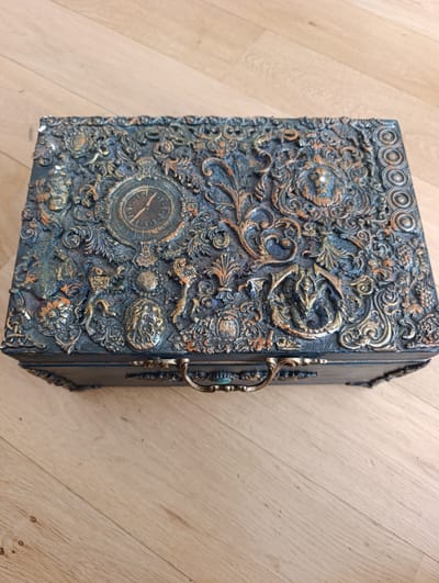 A dark, ornate, rectangular wooden box is displayed on a light-colored wooden floor. The box is covered in intricate, three-dimensional decorative elements, painted in dark blues, bronzes, and oranges, with some elements appearing to be sculpted or applied. Prominently featured on the lid is a decorative clock face with Roman numerals. Other decorative elements include what appear to be lions' heads, swirling scrollwork, and clusters of what might be grapes or berries on the right side. A decorative metal handle is attached to the front of the box. The overall aesthetic of the box suggests a vintage, perhaps steampunk or gothic, style. The lighting is bright, casting subtle shadows, and the scene appears to be indoors, likely on a floor in a room. No people are visible in the image. There is no visible text. The context of Toruń, Poland, is provided but not visually evident in the image itself.