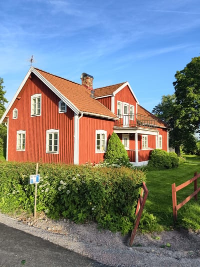 The image shows a bright red, traditional Swedish wooden house with a terracotta tiled roof, located in Slaka, Sweden. The house has white trim around its windows and doors. A balcony with a black railing is visible on the second floor, accessed by a white door. The house is surrounded by lush green grass and dense green hedges. A small, blue sign with white icons is planted on a wooden post in front of the hedges. A broken red wooden fence is partially visible in the foreground, leaning precariously. The sky is a clear, vibrant blue with a few wispy white clouds. A TV antenna is mounted on the peak of the roof, and a brick chimney rises from the upper section of the house. The image appears to have been taken on a sunny day, highlighting the vivid colors of the house and its natural surroundings.