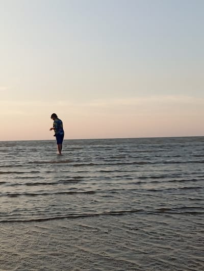 A solitary individual is observed standing in shallow ocean water at a coastal location in Tritunggal, Indonesia. The scene is illuminated by the soft light of either sunrise or sunset. The upper two-thirds of the image show a clear sky, gradating from light blue overhead to a pale orange-pink at the horizon line. The lower third features dark grey-blue water with a rippled surface, indicating calm conditions. The individual, viewed from the back-left, possesses dark hair and is clad in a patterned short-sleeved shirt and dark shorts that extend to just above the knees. The water level reaches approximately the subject's mid-calves. The individual's head is bowed, directing their gaze downward towards the water immediately in front of them. No other persons or significant objects are visible.