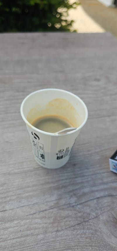 This image, located in Bain-de-Bretagne, France, presents a close-up, high-angle view of a partially consumed beverage in a disposable cup, resting on a wooden surface.

The central subject is a white, octagonal paper cup, approximately half full with a dark liquid exhibiting a light brown crema layer on its surface, consistent with espresso or black coffee. The interior walls of the cup show light brown staining from the liquid. A small, white, flat stirring stick is partially submerged in the liquid and leans against the cup's rim. The exterior of the cup features various black printed symbols and text, including a recycling symbol and a 'do not litter' icon.

The cup rests on a light grey-brown wooden surface, characterized by distinct horizontal grain patterns. This surface occupies the lower two-thirds of the frame. In the lower right corner, a small, dark-colored rectangular box, with visible blue and white accents, is partially visible.

The background, comprising the upper third of the image, is heavily blurred due to a shallow depth of field. It depicts out-of-focus green foliage and bright, indistinct shapes, suggesting an outdoor setting or view through a window. No individuals or specific actions are visible in the scene.