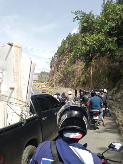 The image depicts a traffic jam on a sunny day, likely in San Antonio de Los Altos, Venezuela. The scene is dominated by a long line of motorcycles and a black pickup truck loaded with a large mattress and other items. The motorcycles are stopped on a paved road that winds through a mountainous, rocky terrain with lush green trees and vegetation lining the sides.

Several people are visible, primarily on the motorcycles, wearing helmets and casual clothing. They appear to be waiting patiently in the traffic. The pickup truck, positioned in the foreground to the left, is packed with goods, suggesting a move or transport of bulky items. The road has yellow dividing lines and a yellow guardrail on the right side. In the distance, more vehicles and people can be seen, along with buildings and a clear sky with some clouds. The overall atmosphere is that of a typical day with slow-moving traffic in a scenic, somewhat rugged environment. The time of day appears to be late morning or early afternoon, given the bright sunlight and clear visibility.

Visible text includes the brand name "TOYOTA" on the red lining of a helmet in the foreground, and a license plate on a motorcycle in the background which reads "ACJ586L".