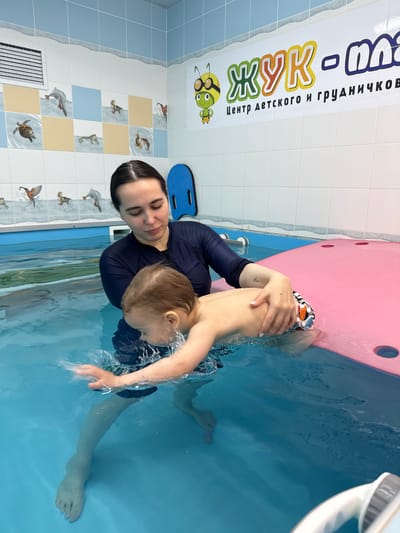 A woman and a young child are in a bright blue swimming pool, likely during a swimming lesson. The woman, wearing a dark long-sleeved swim top, is holding the child, who is wearing swim trunks, as they move through the water, creating splashes. The woman has dark hair pulled back and appears focused on the child. The child's face is partially obscured by water and their position.

The setting is an indoor pool with light-colored tiled walls. The lower section of the walls features a pattern of light blue and light yellow squares interspersed with white tiles, many of which are decorated with images of various birds (ducks, a kingfisher) and a turtle, suggesting a child-friendly environment. A blue kickboard rests on the pool deck to the left of the woman. A pink foam float or mat is visible along the edge of the pool behind the child.

Above the decorative tiles, a white banner with colorful text in Russian is displayed. The main text reads "ЖУК - МАЛ" (Zhuk - Mal), which translates to "Beetle - Small" or "Bug - Little." Below this, smaller text reads "Центр детского и грудничкового плавания," meaning "Center for Children's and Infant Swimming." A cartoon bee or bug wearing goggles and a crown is part of the logo on the banner. The water in the pool is clear and blue.