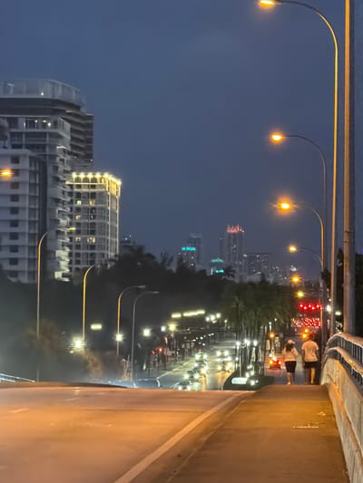 This is a photograph taken at dusk or night on a bridge in Bal Harbour Village, United States of America, overlooking a cityscape and a busy street. The foreground features a concrete bridge with a walkway. Two individuals, a man and a woman, are walking away from the camera on the walkway. The man is wearing a white t-shirt and shorts, and the woman is wearing a white cap, a white top, and a dark skirt.
In the mid-ground, there is a street with multiple lanes of traffic, illuminated by streetlights and the headlights and taillights of vehicles. A traffic light showing red is visible. Palm trees line the street. In the background, a city skyline is visible with several tall buildings, some of which are illuminated. One prominent building appears to be under construction. The sky is dark blue, indicating nighttime. The overall atmosphere is urban and illuminated.