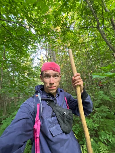 A person is taking a selfie in a forest. The person is wearing a blue jacket, a pink bandana, and a fanny pack. They are holding a wooden stick. The forest is green and lush with trees and leaves. The time of day appears to be daytime with sunlight filtering through the trees. The person's expression is neutral, and they appear to be on an outdoor excursion. The bandana has red lettering on it that reads "I'll be in the". The location is Badashka, Russia.