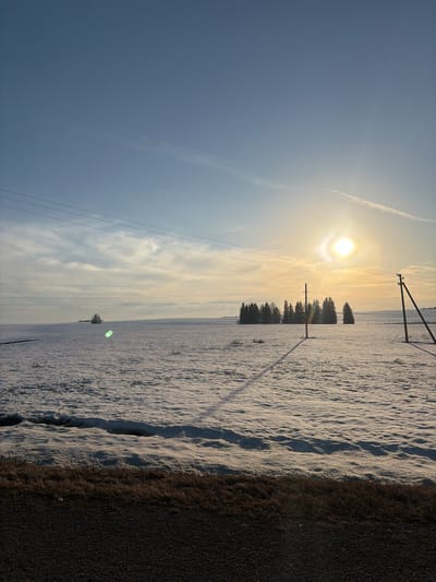 The image depicts a winter scene, likely in the vicinity of Bol'shaya Sosnova, Russia, given the presence of snowy fields and characteristic power lines. The scene shows a field covered in snow. The sun is low on the horizon, partially obscured by light clouds, casting long shadows from the power lines and some trees. A road or path with dark material is visible on the right side of the frame. There are no people visible in the frame. The sky is a gradient of blue to a brighter yellow near the sun. A contrail from an aircraft is also visible.