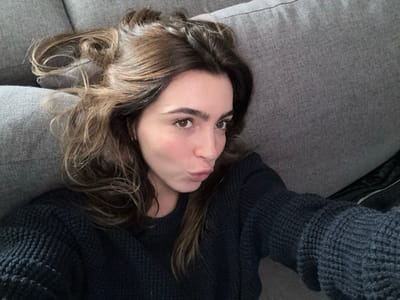 A single female individual is depicted in an indoor setting located in Nogueira e Silva Escura, Portugal. The subject is observed lying supine on a textured gray fabric sofa, her head positioned towards the upper left of the frame. She has long, wavy brown hair and brown eyes. Her face is directed towards the camera, with lips pursed into a pouting formation. She is attired in a dark blue or gray knitted sweater. A dark, possibly black, object resembling a garment or bag is partially visible in the lower right background, resting on the sofa.