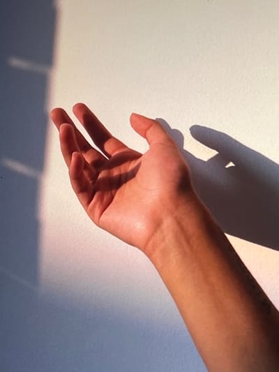 A close-up, eye-level shot features a person's right arm and hand extended toward a white wall bathed in warm, angled sunlight, casting a distinct shadow. The hand is open, palm facing away from the viewer, with fingers slightly curled. The skin tone of the arm and hand appears to be a warm, tanned hue. The wall exhibits subtle texture and is partially illuminated by the sunlight, creating a gradient of light and shadow. On the left side of the image, a darker shadow falls across the wall, originating from an unseen object. A faint pinkish glow is visible at the edge of the shadow on the wall, suggesting the quality of the light. The background is the white wall, with some slight variations in tone due to the lighting. No specific location in Genemuiden, Netherlands, can be identified from the provided image.