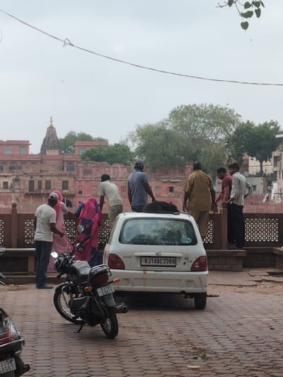 The image depicts a street scene in Jodhpur, India, with a temple structure visible in the background. In the foreground, a white car is parked, and a black dog is lying on its roof. Several people are standing around the car and on a raised platform behind it. A motorcycle is parked to the left of the car. The ground is paved with hexagonal stones. The sky is overcast, suggesting a cloudy day. The architecture in the background, characterized by its red sandstone construction and ornate detailing, is consistent with the style found in Jodhpur.