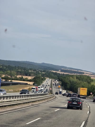 The image shows a view of a highway in Würges, Germany, with a line of traffic moving in the foreground and background. The sky is overcast with a light blue-gray hue. The highway has multiple lanes, separated by guardrails and white lines. In the foreground, a black BMW sedan with a German license plate "MTK SE 821" is driving on the right side of the road. Several other vehicles, including cars and trucks, are visible on the highway, some in traffic. The landscape to the left of the highway features a solar panel installation and fields of dry grass or crops, followed by rolling hills covered with trees. To the right, the landscape is also wooded with trees. Speed limit signs of "120" are visible on the highway.