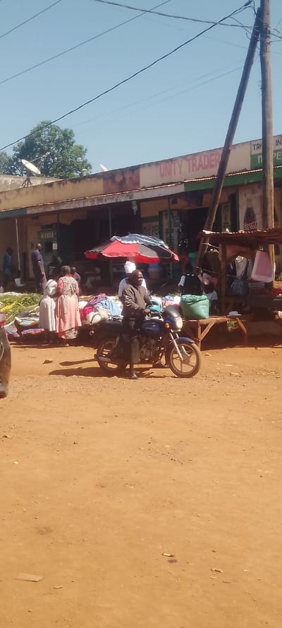 The image displays an outdoor market scene in Chemuche ward, Kenya, under a clear blue sky with overhead power lines. The ground is unpaved reddish-brown dirt.
In the foreground, slightly right of center, a man wearing a brown jacket and dark trousers is seated on a dark blue motorcycle, facing right with his left foot on the ground. A partial view of a dark vehicle bumper is visible on the far left.
The midground features an active open-air market. Two women, one in a light pink patterned dress and another in a cream-colored garment, stand near ground-level displays of fabrics and goods to the left of the motorcycle. A large red and black striped umbrella shades a market stall area laden with various merchandise. To the right, a wooden market stall with a basic roof structure exhibits hanging clothes and a green sack. Several other individuals are dispersed throughout this area, some standing, suggesting commercial interactions.
The background is dominated by a single-story building with "UNITY TRADER" painted in red on its upper facade. This building has a corrugated metal roof supporting two white satellite dishes. Its open shopfronts display "M-PESA" signage, and a Coca-Cola advertisement is visible on the right section. A tall wooden utility pole stands to the right of the building. Green tree foliage is visible above and behind the building, to the left. The scene is brightly lit by direct sunlight, casting noticeable shadows.