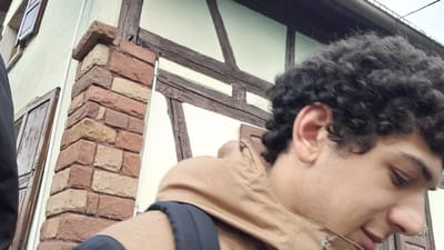 An individual is depicted in the foreground, positioned on the right side of the frame, against the backdrop of a building in Meistratzheim, France. The subject, a male with dark, curly hair, is seen in profile, looking downwards and to the right. He is wearing a light brown hooded jacket, with a dark strap, likely from a backpack, visible over his right shoulder.
The background structure displays architectural elements characteristic of the region. A section of a light-colored facade with a window featuring open dark brown wooden shutters is visible on the left. This transitions to a prominent corner pillar constructed from reddish-brown and beige rough-cut stone or brick blocks, with a thin white pipe running vertically alongside it. Beyond this, a half-timbered section of the building is visible, featuring dark brown wooden beams set against a light-colored infill. The upper background indicates an overcast sky, and faint streaks consistent with light precipitation are present. No other individuals or discernible interactions are depicted.