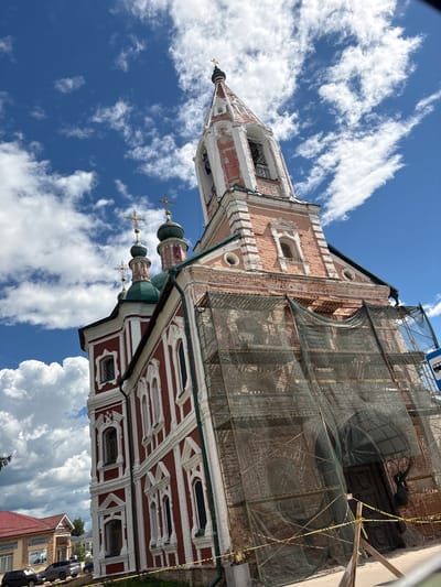 The image depicts the Church of John the Warrior in Pereslavl-Zalessky, Russia, on a bright, cloudy day. The church is an Orthodox Christian structure with red and white facades, multiple green domes topped with crosses, and a prominent bell tower. The bell tower is partially covered in scaffolding and netting, suggesting ongoing renovations. In the foreground, construction barriers and caution tape are visible. The background shows a clear blue sky with scattered white clouds and a few trees. In the lower left corner, a building with signs in Russian and parked cars can be seen.