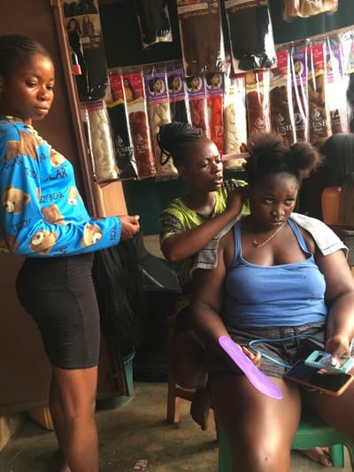 The image depicts the interior of what appears to be a hair salon or beauty supply store in Isieke, Nigeria. There are three African women in the frame. One woman is lying down with her head resting near a display of packaged hair extensions, which are arranged on a shelf. Another woman is sitting down, having her hair styled by another woman who is standing behind her. The seated woman is wearing a blue tank top and is holding a purple comb and a smartphone. The woman styling her hair is wearing a green and yellow patterned top. The environment includes products for hair styling, chairs, and a hint of the exterior visible in the background.