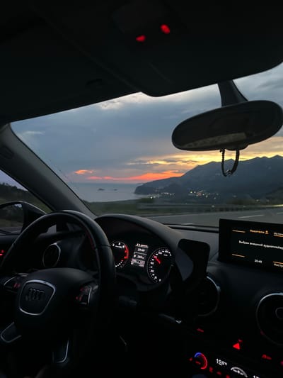The image captures the interior of a car driving along a coastal road at dusk, with a view of the Adriatic Sea and mountains in the distance. The dashboard displays the time as 20:49 and the speed as 160 km/h. The setting is likely Kanjoši, Montenegro, given the characteristic coastal scenery and mountainous terrain. The sky is painted with hues of orange, pink, and blue from the setting sun, casting a warm glow over the landscape. Lights from a town are visible nestled on the mountainside, adding a touch of human presence to the natural beauty. The car's interior is dimly lit, with the dashboard lights and the sunset providing the primary illumination. The steering wheel and other car components are visible, suggesting the perspective of a driver or passenger. The overall atmosphere is one of serene travel, with the beauty of nature unfolding outside the car.