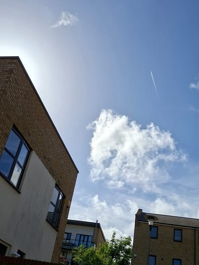 The image presents an upward-looking, dynamic view of modern buildings against a blue sky with clouds, taken in Dartford, United Kingdom. The scene is dominated by architectural elements and the expansive sky.

In the foreground, on the left side, a multi-story brick building with dark-framed, reflective windows slants across the lower and middle sections of the frame. Part of its facade is a contrasting lighter, possibly rendered, material. A strong glare from the sun is visible in the lower-left corner, casting brightness onto the building and sky.

On the upper right, another brick building, featuring similar dark-framed windows and a dark roof line, is partially visible. A black street light or lamp post with a circular fixture protrudes from this building's side. Below this structure, a third building is partially obscured by vibrant green foliage, likely from a nearby tree.

The sky, filling a significant portion of the image, is a clear, bright blue. It features scattered white, cumulus-like clouds, with a particularly large and well-defined cloud formation visible on the right. A thin, white contrail from an airplane stretches diagonally across the upper-left quadrant of the sky, indicating high-altitude air travel.

The perspective is taken from ground level, looking up, emphasizing the height and angularity of the buildings. The bright conditions suggest a clear, sunny daytime. No people or visible text are present in the image.