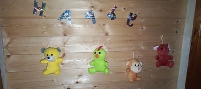Four small plush toys are hanging by strings against a wooden wall. From left to right, the toys are a yellow bear, a green chick with an orange beak, an orange monkey, and a red elephant. Above the toys, a banner made of shiny, holographic letters spells out "HAPPY". The wooden wall has a natural, unfinished appearance with visible grain and knots. The scene is indoors, likely a room or a covered area, as indicated by the artificial lighting and the presence of a decorative banner. The location of Мерчалово Снт, Russia is noted for context, but the image itself provides no specific visual cues to confirm this location beyond the general description of the setting.