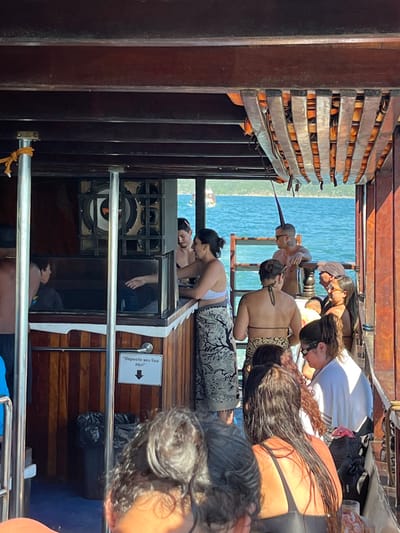 People are on a wooden boat. Several people are looking over the edge toward the water. Others are standing or sitting nearby. A speaker is visible. A sign on the wood paneling says, "Deposite seu lixo aqui" with an arrow pointing to the left. The sea can be seen in the background.