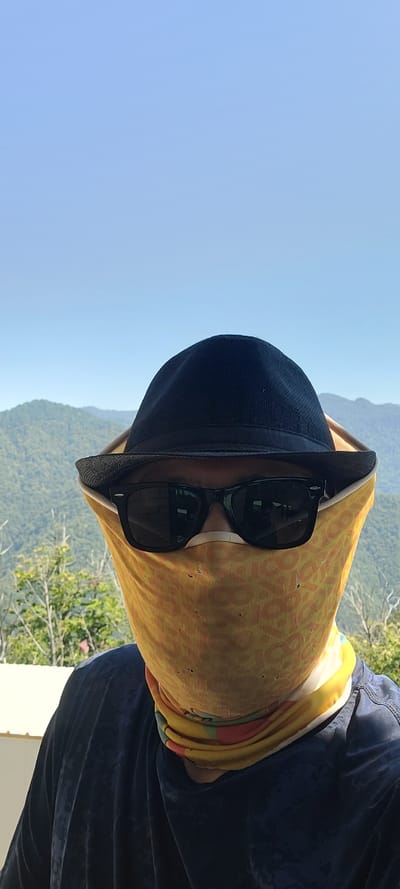 In Milisi, Georgia, a person is photographed outdoors with a backdrop of lush green mountains under a clear blue sky. The individual is wearing a black fedora hat, sunglasses, and a yellow face covering patterned with an orange design. Their shoulders are visible, draped in a dark blue t-shirt with a subtle pattern. The person appears to be looking towards the camera, with the sun casting a slight glare on their sunglasses. The image composition is a close-up, focusing on the person's upper body and face.