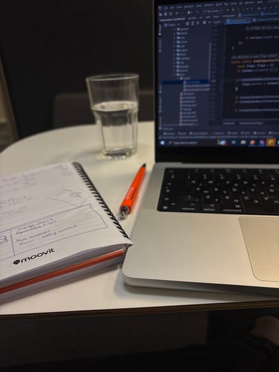 An overhead, slightly angled view captures a workspace setup on a white table with a dark wooden edge, located in Nes Ziona, Israel. A silver MacBook laptop occupies the right side of the frame, open and displaying what appears to be lines of code within an integrated development environment (IDE) on its screen. The laptop's keyboard and trackpad are visible. To the left of the laptop, an orange pen rests on the table, positioned between the laptop and a spiral-bound notebook. The notebook is open to a page featuring handwritten notes, diagrams, and text, including phrases such as "merge flows," "error," and "adding comment." The "Moovit" logo is prominently displayed at the bottom of the open notebook page. Further to the left and slightly behind the notebook and pen, a clear glass partially filled with water sits on the table. The background is out of focus and dark, suggesting an indoor office or cubicle environment. No individuals are present in the image.